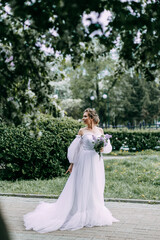 Fototapeta na wymiar A beautiful gentle attractive young woman in a wedding dress with a bouquet of lilacs walks alone and poses in a spring garden with flowering plants