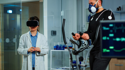 Scientist doctor in sport science laboratory wearing virtual reality goggles while athlete running with electrodes attached on body monitoring physical endurancewhile ekg scan runs on computer screen.