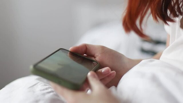 Female hand holding smartphone and watching video on a bed inside home indoors