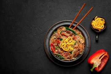 Japchae in black bowl on dark slate table top. Korean cuisine glass chapchae noodles dish with vegetables and meat. Asian traditional food. Authentic meal. Top view. Copy space