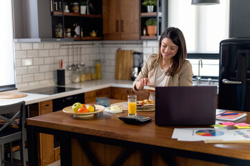 Young smiling business woman working from home sitting in front of laptop computer at home kitchen. Using smart phone