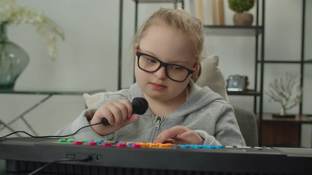 Engrossed in action adorable elementary age girl in eyeglasses with general learning disability singing into microphone, playing on synthesizer, developing music ability and motor skills indoors.