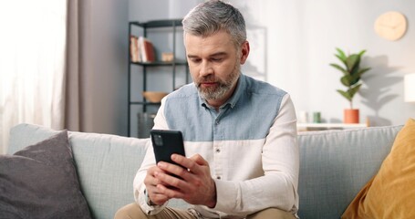 Close up of cheerful handsome middle-aged Caucasian man in positive mood texting on smartphone while sitting at modern home on sofa and smiling, grey-haired male using social network app on cellphone