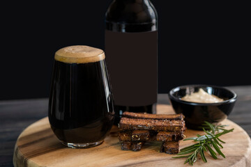 Bottle of dark beer, full beer glass with foam, black bread crackers, gravy in a saucepan, and a sprig of rosemary close-up on a wooden tray