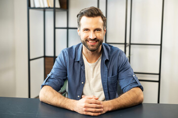 Portrait of smiling businessman with bristle and charming smile, sitting at the desk at modern office, dressed in white t-shirt, denim shirt, freelancer, owner of a new startup, successful leader