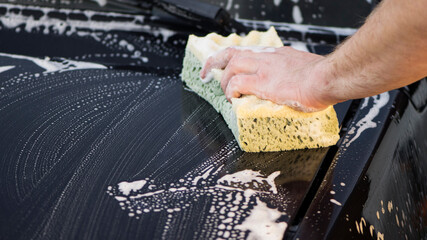 a hand with a washcloth and soap suds washes the hood of the car. Man washing car hood with washcloth and shampoo, auto covered with foam, car wash service. cleaning the black surface