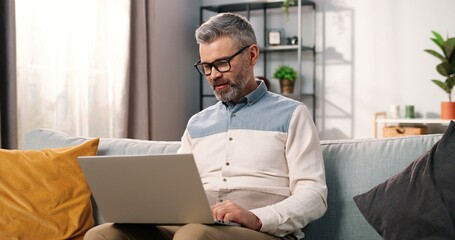 Portrait of joyful Caucasian handsome middle-aged bearded man in glasses sitting on sofa in living room in apartment speaking on video call online on laptop and waving hand, video chat concept