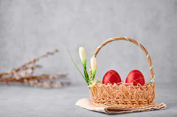 Two red painted Easter eggs in the wicker basket on gray table top with pussy willow branches on backdrop