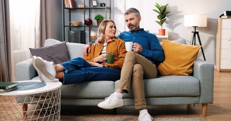 Middle-aged cute lovely Caucasian happy married family couple wife and husband resting on sofa in modern living room at home drinking coffee, sipping hot drink and speaking, love concept