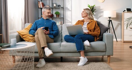 Caucasian married couple resting on sofa in room in apartment using digital gadgets and chatting. Middle-aged man browsing on smartphone while his pretty wife typing on laptop online, leisure concept