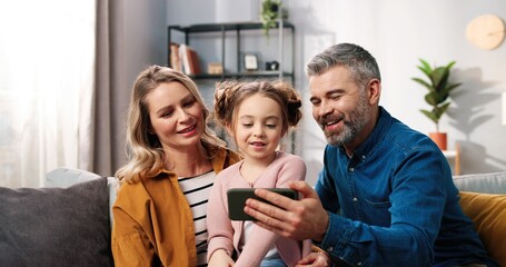 Close up portrait of joyful cute Caucasian family videochatting on smartphone. Happy parents mom and dad with cute small child girl speaking on video call online on cellphone smiling and waving hand