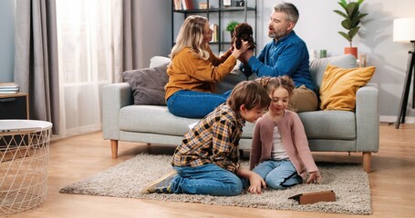 Joyful Caucasian family spending time at home. Happy kids little boy and girl sitting on floor watching videos or cartoons on tablet while parents playing with pet puppy dog on sofa, leisure concept