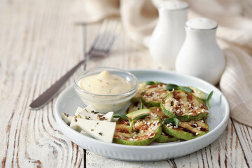 Delicious grilled zucchini slices served with sauce and cheese on white wooden table, closeup