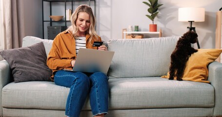 Joyful beautiful Caucasian woman sits on sofa in modern living room with cute puppy dog and buying online making payment with credit card on laptop, e-commerce shopping, sales and discounts