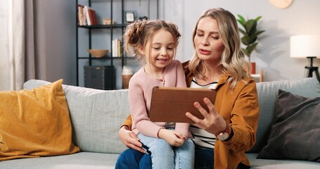 Portrait of happy joyful caring Caucasian mom with small daughter in room together videochatting using tablet. Pretty mother teaching small girl to use gadget device, family time, watching cartoons
