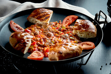 Turkish lamb goulash in red sauce with bell pepper and traditional buns in a metal dish on a black background. Close up, selective focus