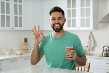 Young man with glass of chocolate milk showing Ok in kitchen