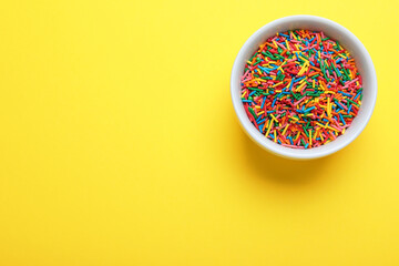 Colorful sprinkles in bowl on yellow background, top view with space for text. Confectionery decor