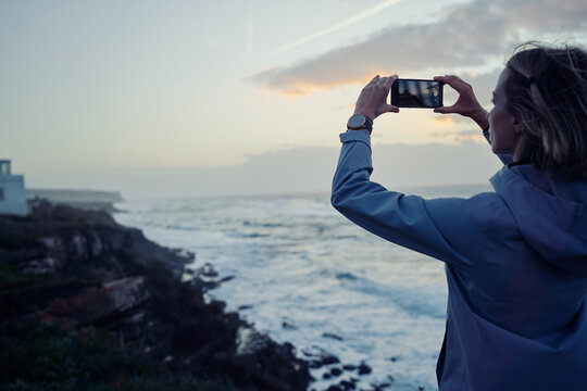 Traveling by Portugal. Young woman taking photo of wonderful sunset on rock ocean shore.