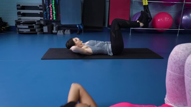 Fit women in sportswear lying on mats with feet resting on TRX straps and doing abdominal exercises. Multiethnic females working out together in gym. Concept of sport
