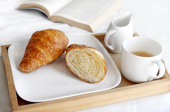Coffee and croissant breakfast tray on a bed next to a book
