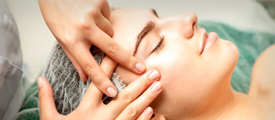 Young caucasian woman receiving facial massage by beautician's hands in spa medical salon