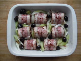 meat rolls in bacon with vegetables, olives and spices in a white plate, on a wooden table, the cooking process.