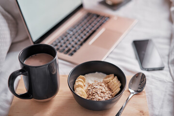 Breakfast in bed: a wooden tray with granola and cup of cocoa with a laptop on background.