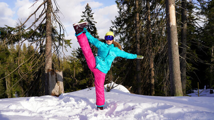 Happy flexible girl snowboarder in bright clothes performs a split in a snowy forest