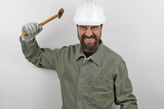 Worker man with beard wearing worker uniform and hardhat holding hammer with angry expression looking at the camera