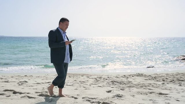 Businessman in elegant suit walking on the sea shore while using smartphone in a sunny day after work searching for a quiet place.