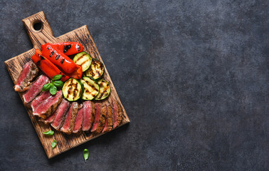 Slices of grilled beef steak and grilled vegetables on a wooden board on a black background, top view.