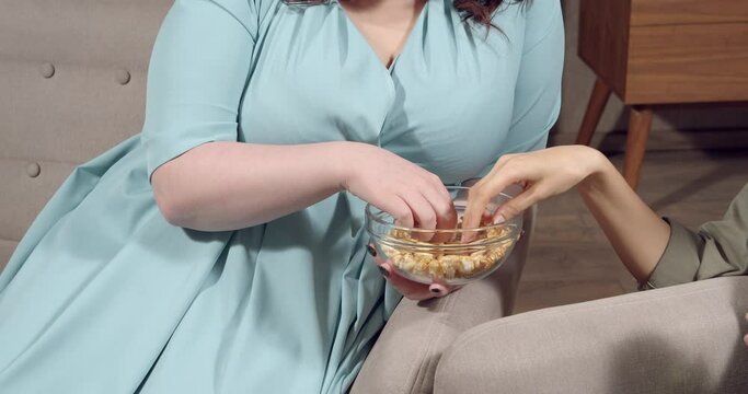 Unrecognizable plus size woman eating popcorn sitting on the couch. Hands of women taking snacks from a bowl while watching a movie at home.