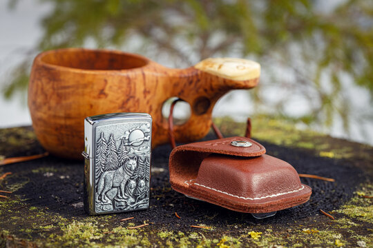 Zippo Timberwolves lighter with a leather case and a wooden kuksa with delicious coffee on an old stump in the spring forest. The background is blurred.