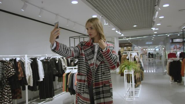 Young woman dressed in plaid coat is taking a selfie at clothing store, tracking shot, 360 degree