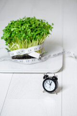 micro-green on the scale for measuring body weight, centimeter tape for the body, clock on a light background. means and items for a diet, a healthy lifestyle concept . vegan superfood trend