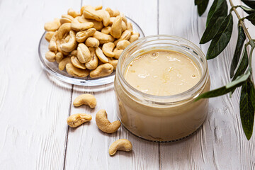 A jar of cashew butter with a bunch of fresh cashew on a white wooden table with an olive branch. Homemade cashew butter, natural, healthy food. The modern wellness and vegan concept.