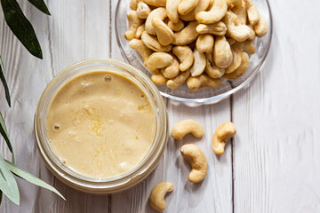 A jar of cashew butter with a bunch of fresh cashew on a white wooden table with an olive branch. Homemade cashew butter, natural, healthy food. The modern wellness and vegan concept.