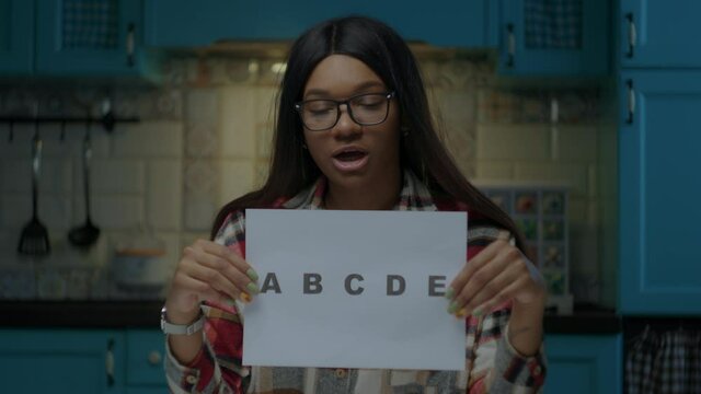 Female African American teacher in glasses talking to camera and showing sheet of paper with ABCDE letters. Webcam view of working from home online school teacher.