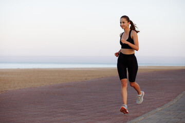 girl runner in a black tracksuit and sneakers runs by the sea during sunset