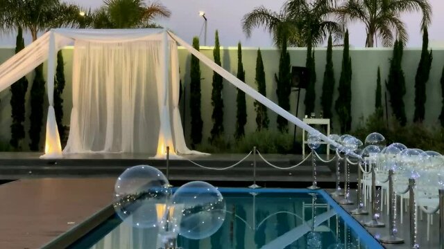 Jewish traditions wedding outdoor ceremony. Wedding canopy (chuppah, hupa) made of white fabric against the background of evening sky, palms and an artificial reservoir decorated with balloons. HD