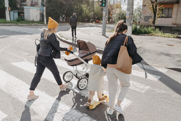 mother transfers a child riding a scooter on a crosswalk, and another woman with a stroller rides next to them