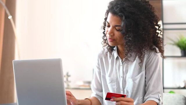 Beautiful biracial young woman holds a debit card and enters data on the laptop, shopping online. A satisfied African girl is making order on the computer, purchasing
