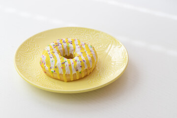 Confectionery product. Donut decorated with yellow-white icing. The donut is on a yellow dessert plate on a white table.