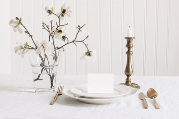 Wedding stationery mock-up. Blank business, RSVP paper card on plate. Festive table setting with brass candleholder. Linen tablecloth background. Blooming white star magnolia tree branches in vase.