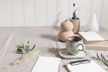 Artistic wokspace, still life. Paint brushes and pencils. Olive branches, vase, cup of coffee and blank paper card mockups on linen tablecloth. Art supplies scene with dry grass. Table background.