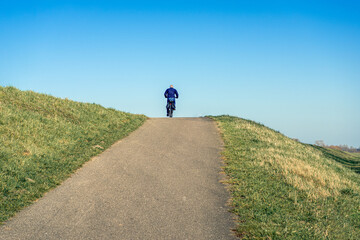Unidentified man cycles on the driveway to the road on top of the Dutch dike. The photo was taken on a sunny day in the winter season with a clear blue sky.