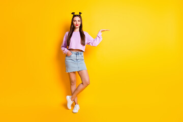 Fototapeta na wymiar Full size photo of optimistic pretty brunette girl hold empty space wear pink sweater skirt sneakers isolated on yellow background