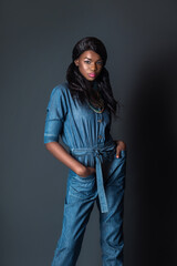 Portrait of an elegant black woman with curly long black hair and beautiful makeup posing by herself in a studio with grey background wearing a denim jumpsuit and colorful jewelry.