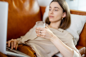 Pretty woman hold a glass of water while working on laptop and lying on sofa. Lifestyle and healthy concept. Dehydration. Rate of drinking water per day. Focus on the glass.